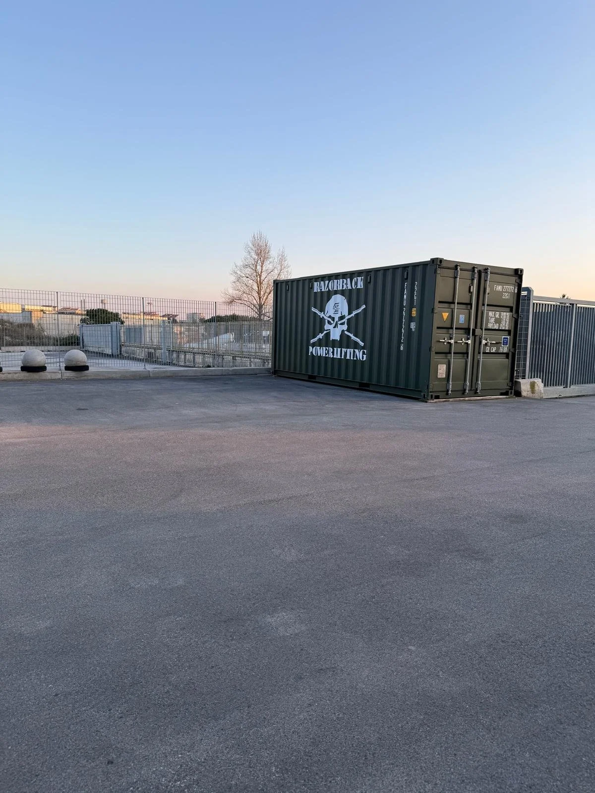Container militare di colore verde con la scritta "TRAZBACK POWERLIFTING" e un logo di un teschio con due bastoni incrociati, posto su una terrazza con parapetto in metallo, sotto un cielo sereno e senza persone visibili.