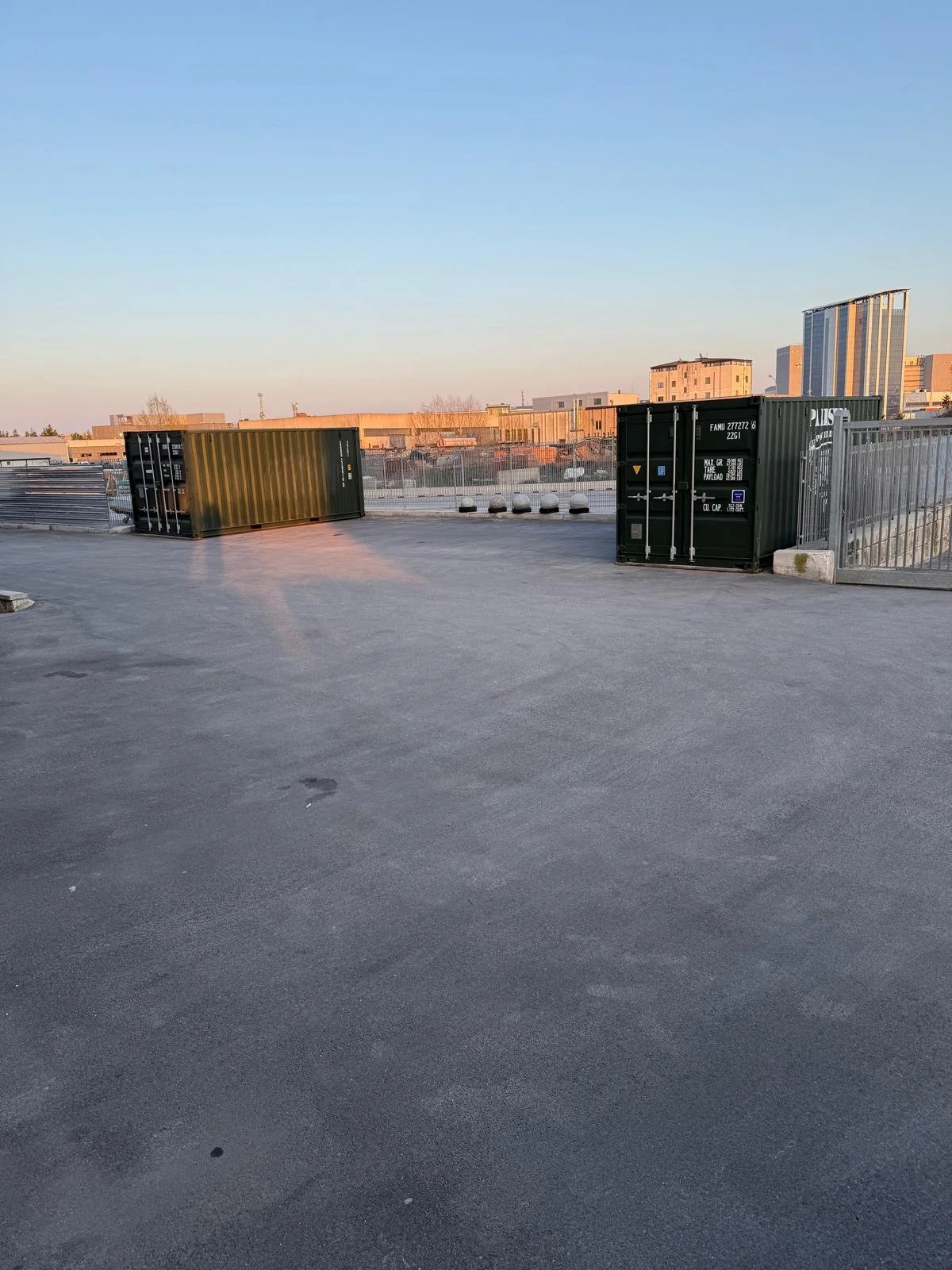 Due container di colore verde su una superficie asfaltata, con un cancello e un elemento di sicurezza in metallo dietro, e un'area urbana con edifici al tramonto sullo sfondo.
