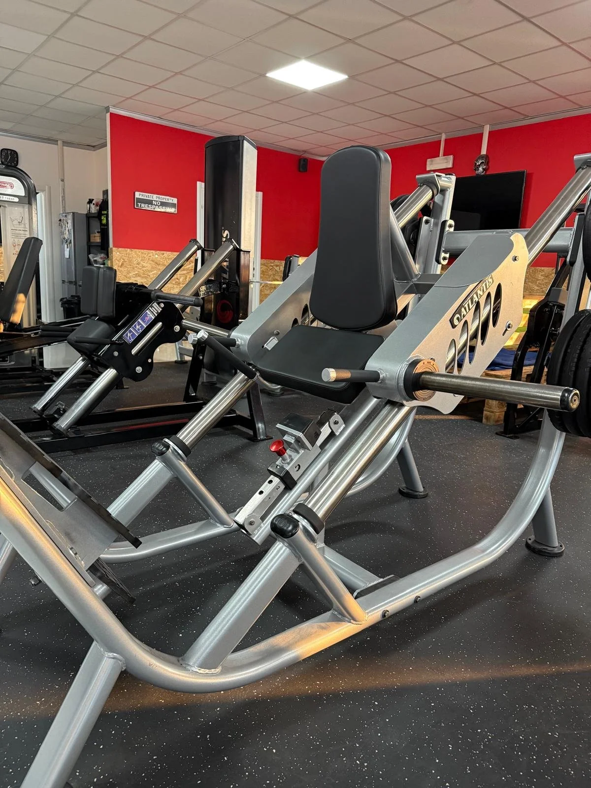 Attrezzi da palestra, tra cui una macchina per affondi e altre attrezzature di allenamento, in una palestra con pareti rosse e un soffitto a pannelli.