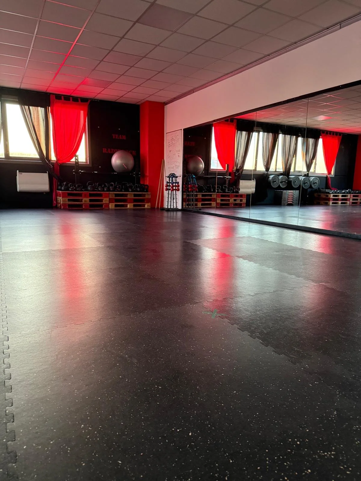 Studio di fitness o palestra vuota con specchio grande, tappeto nero, palloni fitness, kettlebell e barbell, con tende rosse e nere alla finestra.
