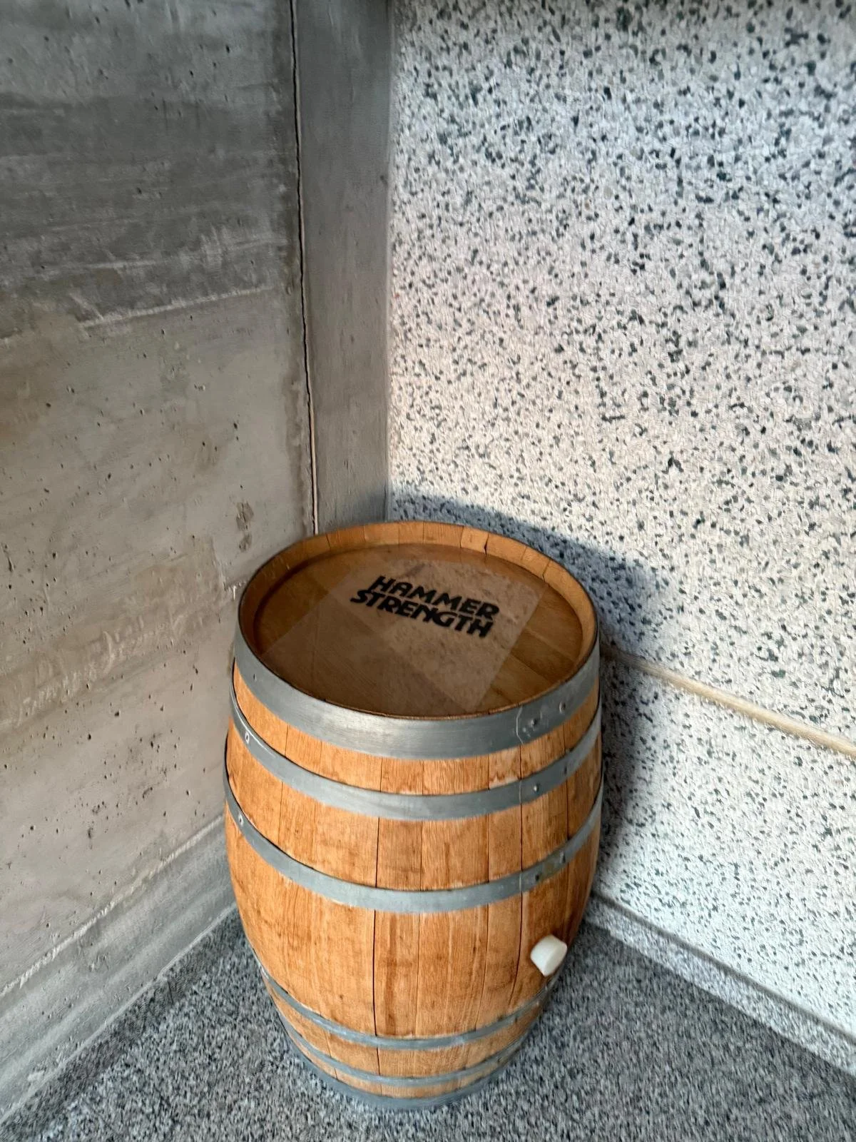 Barile di legno con la scritta 'Hammer Strength' sul coperchio, posizionato in un angolo tra parete di cemento e parete di cemento con finitura a mosaico.