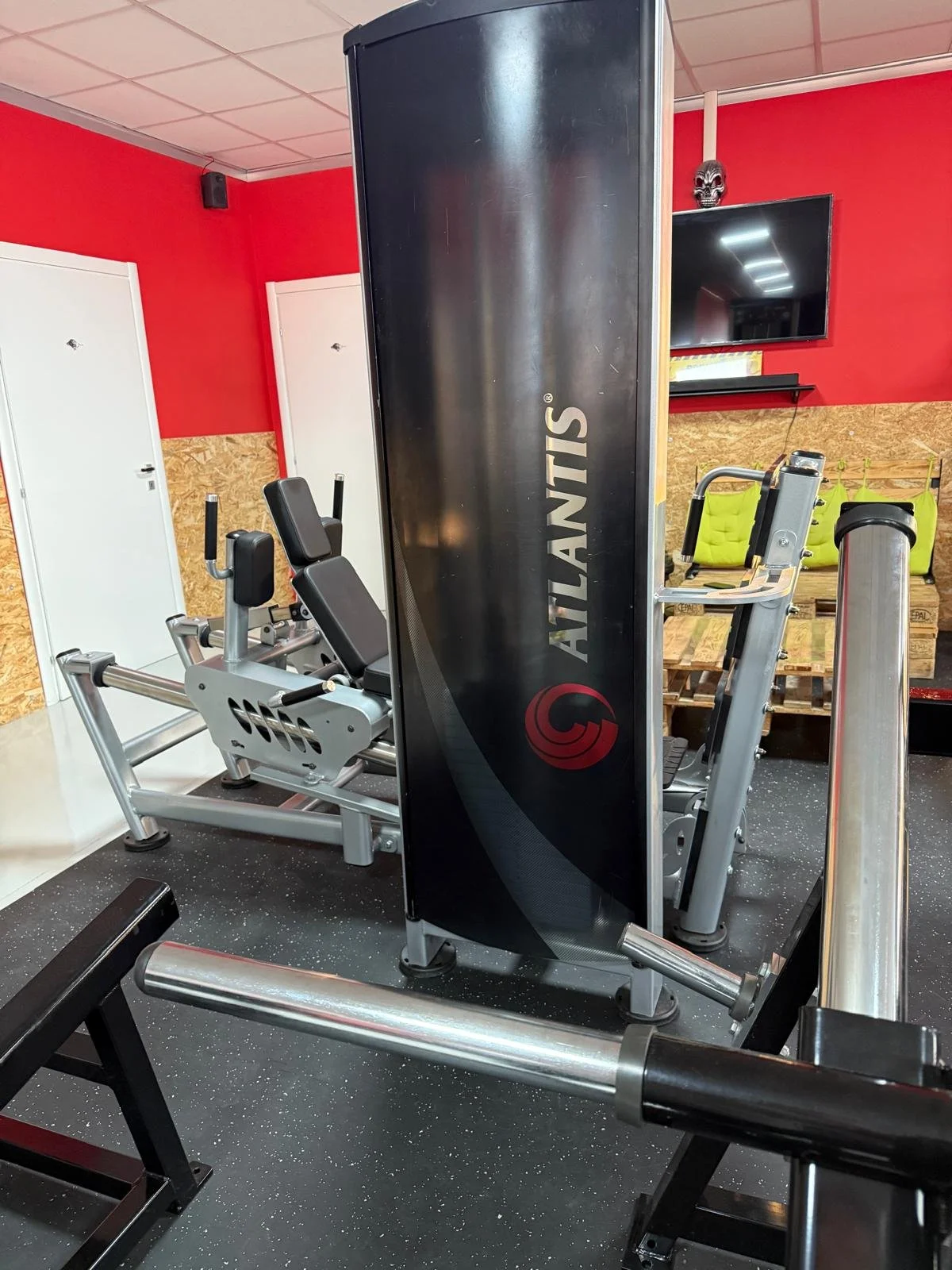 Attrezzi da palestra, compreso una macchina per esercizi e una panca, in uno spazio con pareti rosse e pavimento nero con puntini biancastri, decorazioni come uno skull e un televisore.
