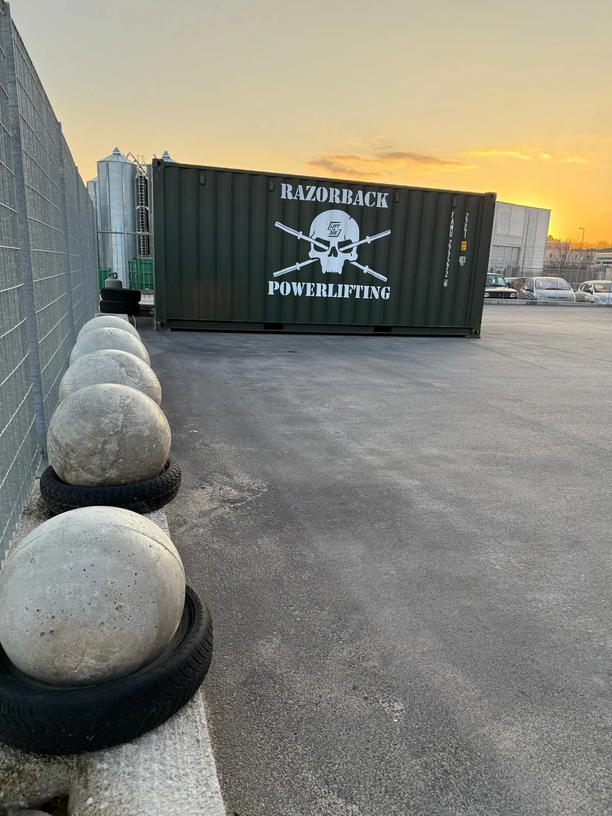 Contenitore di metallo verde con logo di una testa di teschio e frecce incrociate, scritto 'Razorbuck Powerlifting', in un parcheggio con balle di cemento e pneumatici usati ai lati.