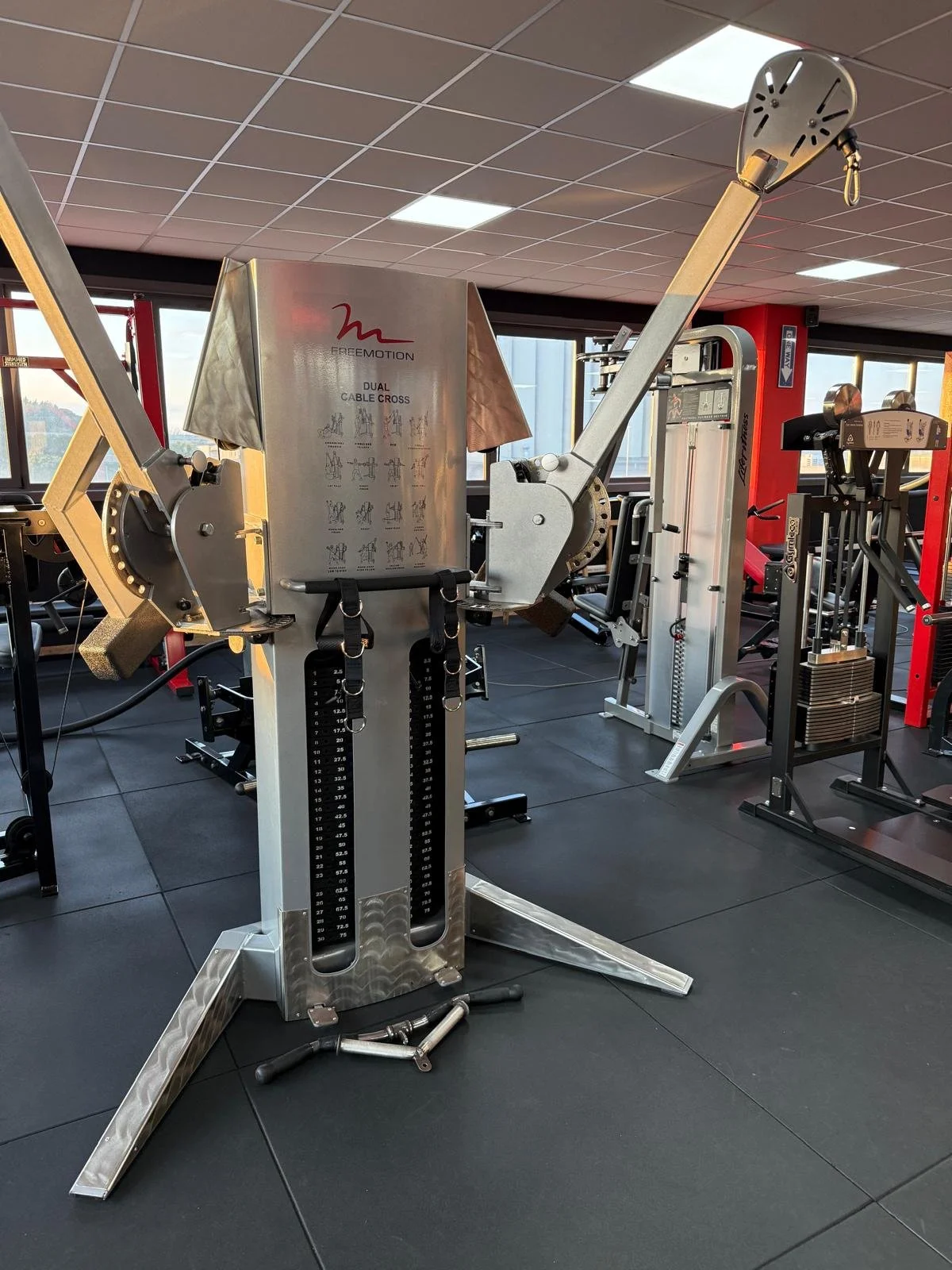 Attrezzi da palestra, inclusa una macchina per trazioni muscolari, in una sala fitness moderna con grandi finestre a vista e pavimento in piastrelle nere.