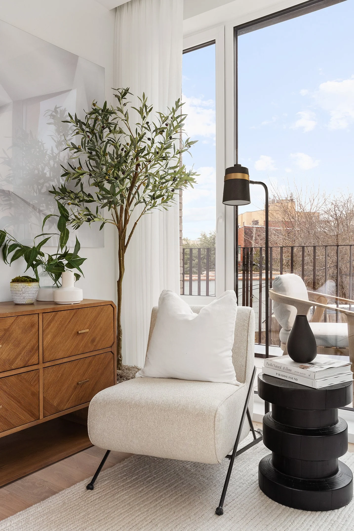 Interior of a bright living room with a beige armchair, a black side table with stacked books and a black vase, a modern floor lamp, wooden sideboard with plants, large glass door leading to a balcony with outdoor chairs, and a view of a sunny sky an