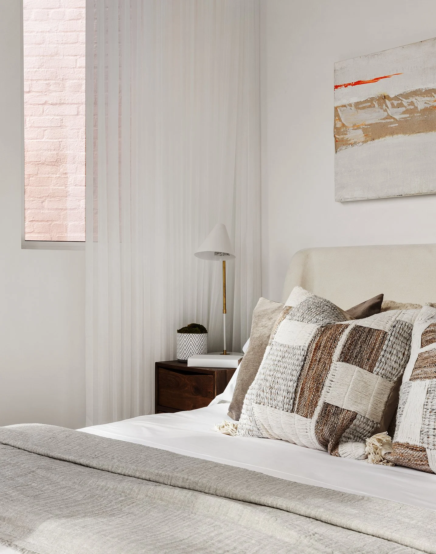 A bedroom with a bed, patterned pillows, a nightstand with a potted plant, a white curtain, and a wall-mounted abstract painting.