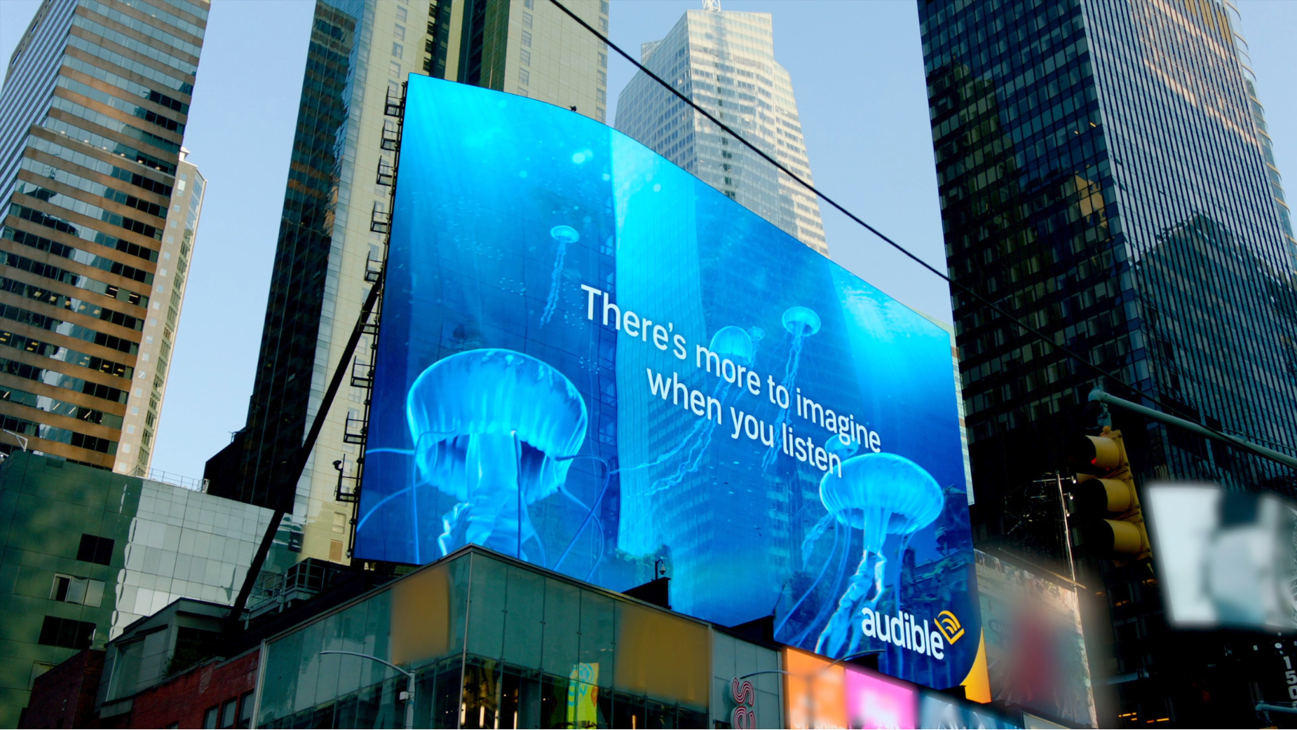 Digital billboard in Times Square displays jellyfish and lightning with the text 'There's more to imagine when you listen' and the Audible logo.