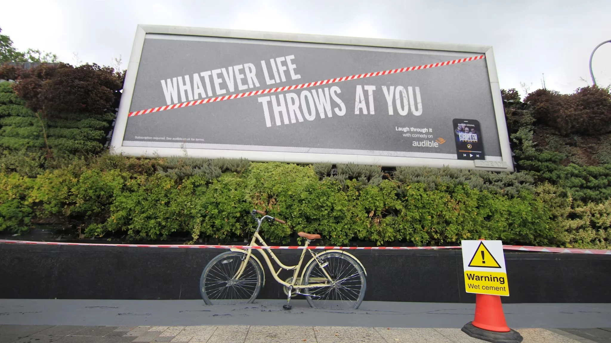 A large billboard with a grey background and white text that reads "Whatever life throws at you". Below the billboard, there is a yellow warning sign that says "Warning Wet cement". A white bicycle with a brown seat is parked in front of the billboar