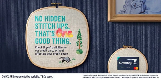 Embroidery hoop with a motivational message about honesty and credit cards, featuring small illustrations of a tree and a weasel, hanging on a blue wall next to a framed photo and a Capital One credit card.