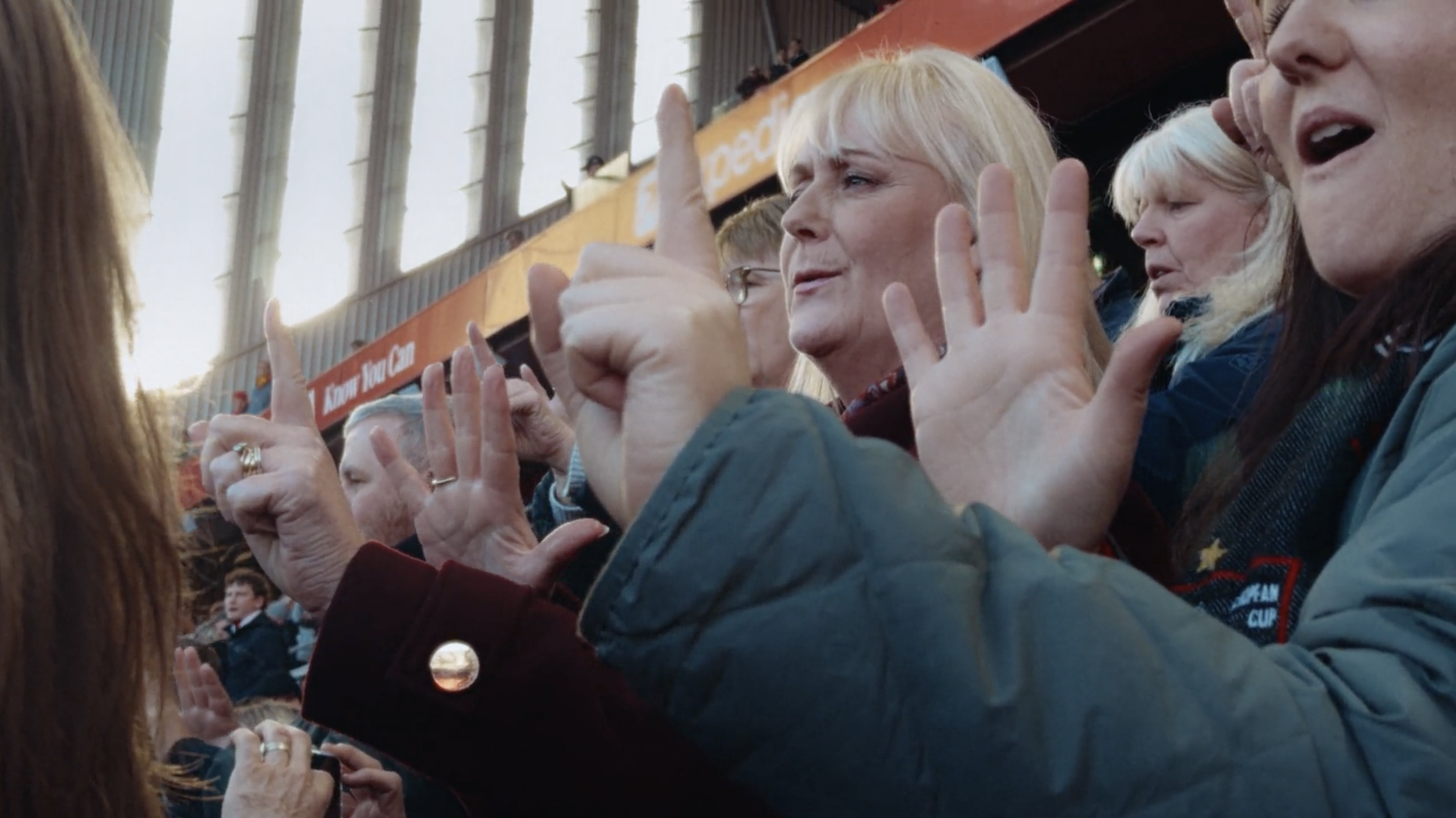 Group of people sitting in a stadium, engaging in conversation and waving, with large windows and bright sunlight in the background.