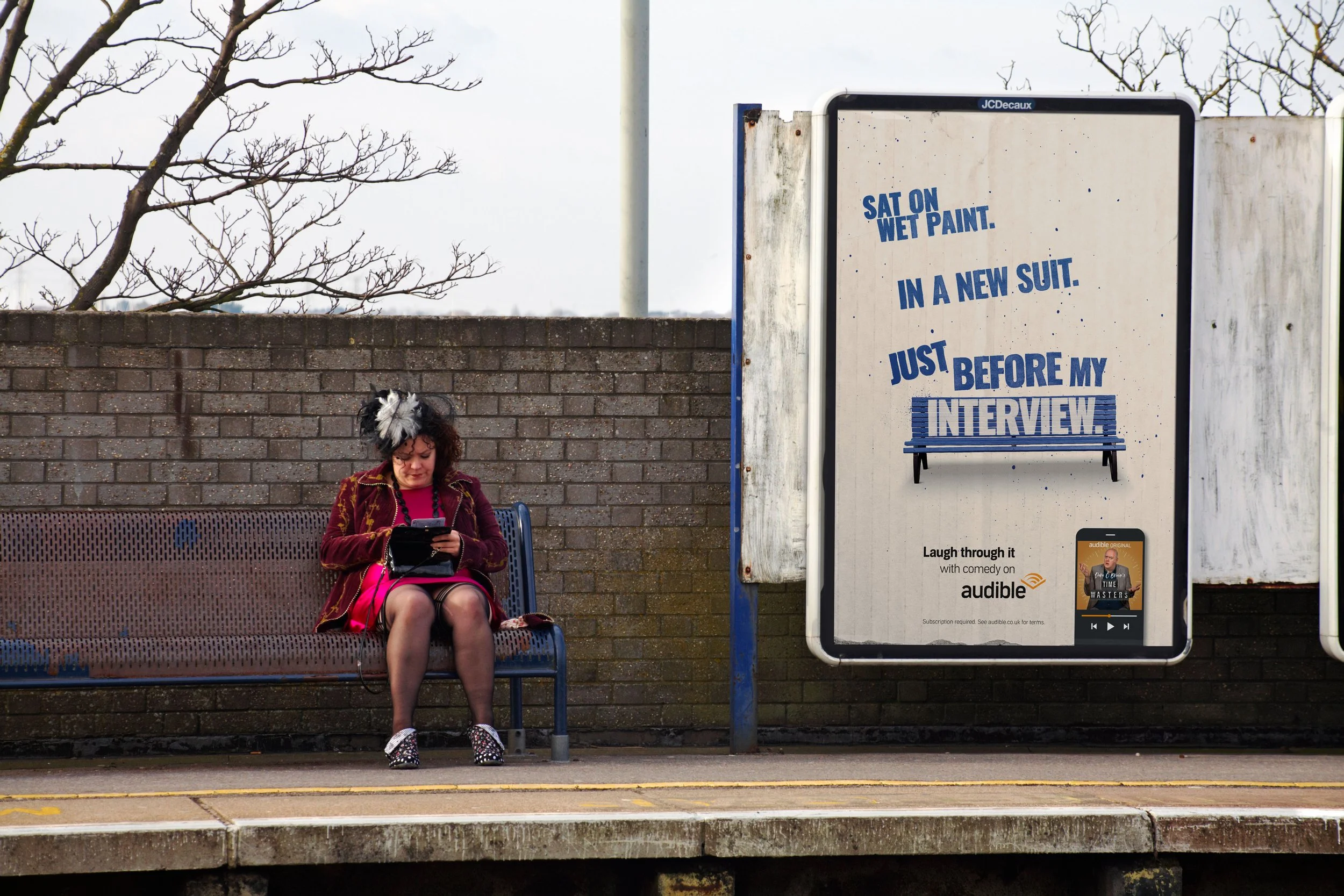 A woman with curly hair wearing a feathered headpiece and a maroon jacket sitting on a bench at a bus stop, looking at her phone. There is a large advertisement board next to her with a bench illustration and promotional text for Audible. A brick wal