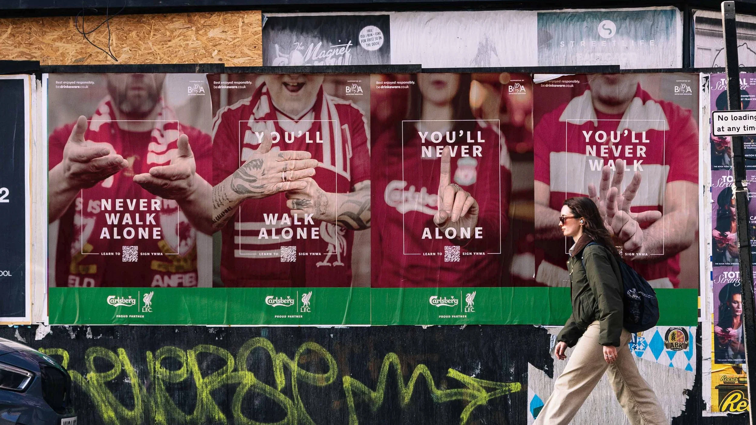 Posters on a wall featuring football players and message 'You'll Never Walk Alone.' A woman crosses the street in front of the posters.
