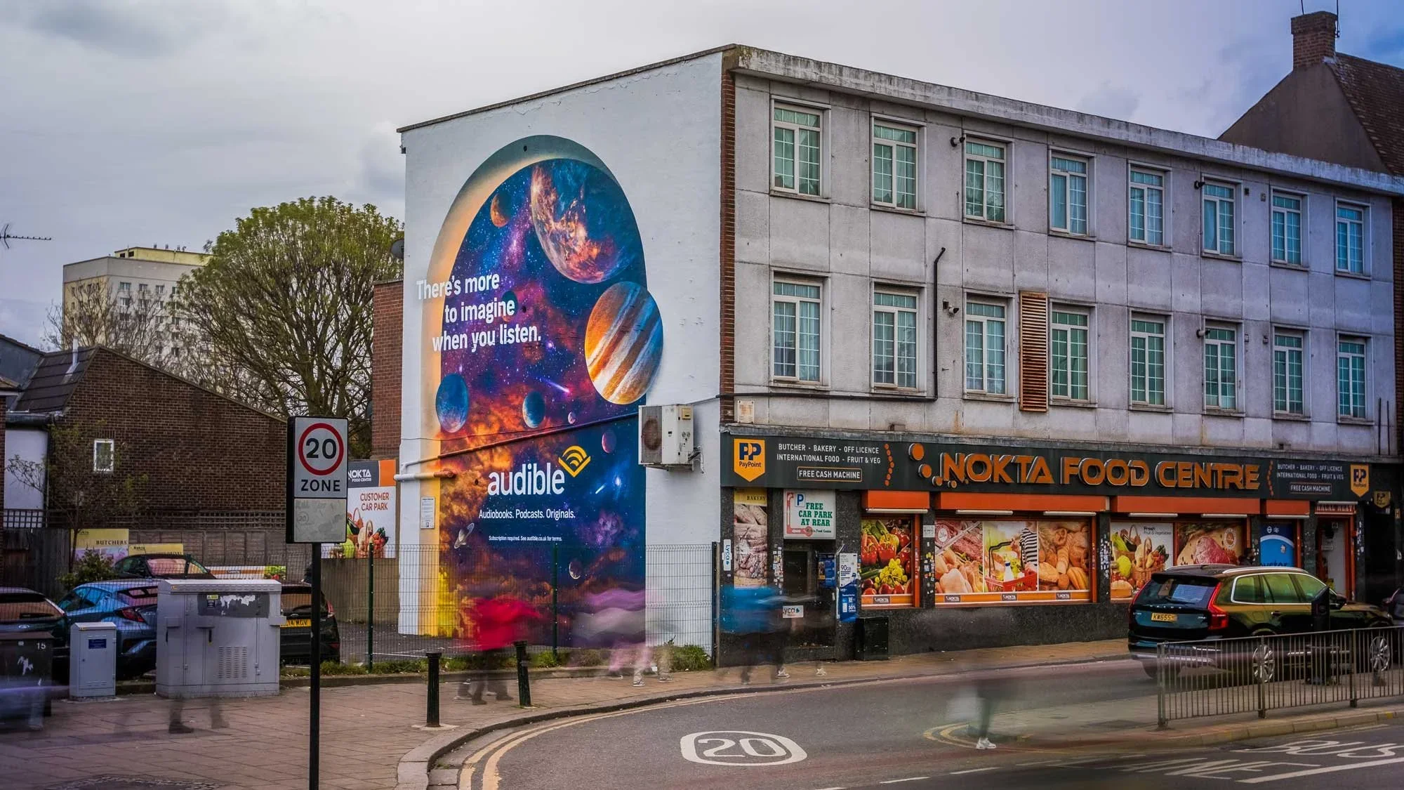 A large mural on the side of a building features a cosmic scene with planets and stars and the text 'There's more to imagine when you listen.' Below the mural, a sign for audible is visible. The building houses Nokta Food Centre, which sells food and