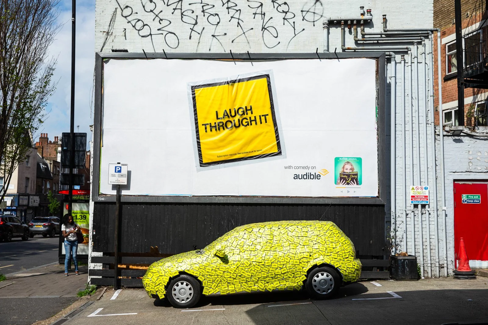 A yellow car covered in numerous yellow sticky notes parked in front of a large white billboard. The billboard displays an advertisement with the text 'Laugh Through It' in bold black letters on a yellow background, along with the phrase 'with comedy