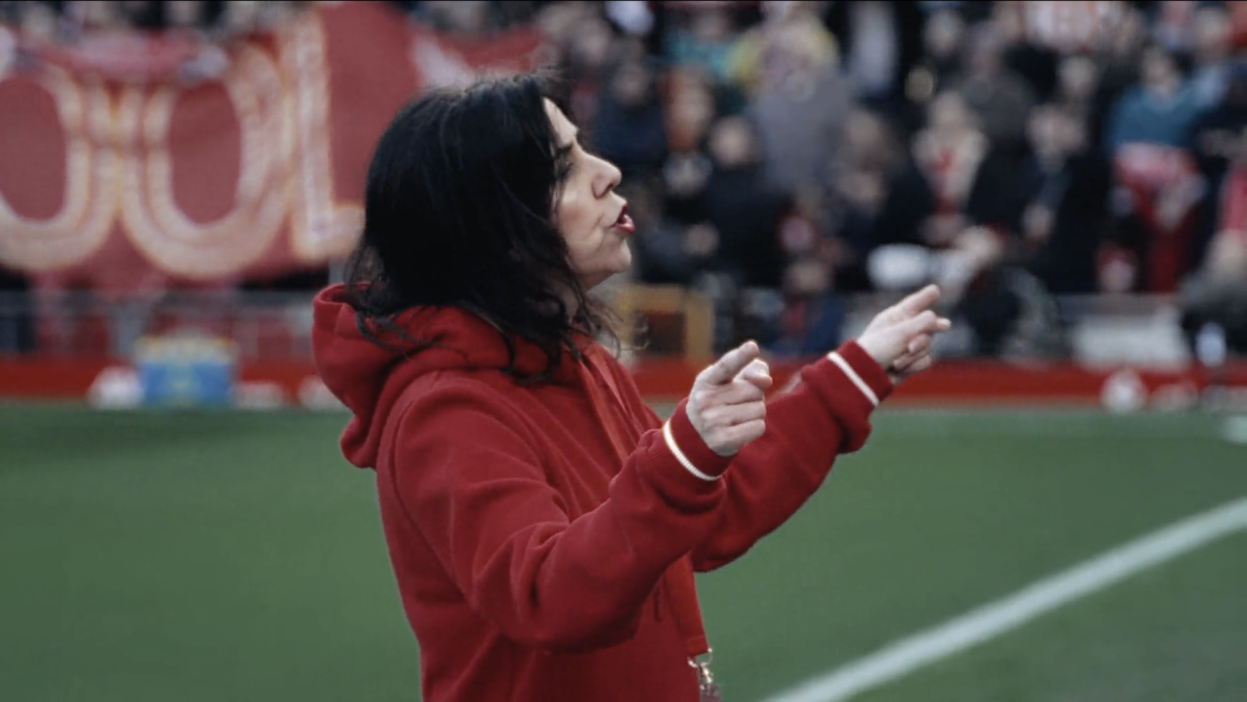 A woman with black hair, wearing a red hoodie, is making a gesture with her hands on a sports field with a blurred crowd and colorful banners in the background.