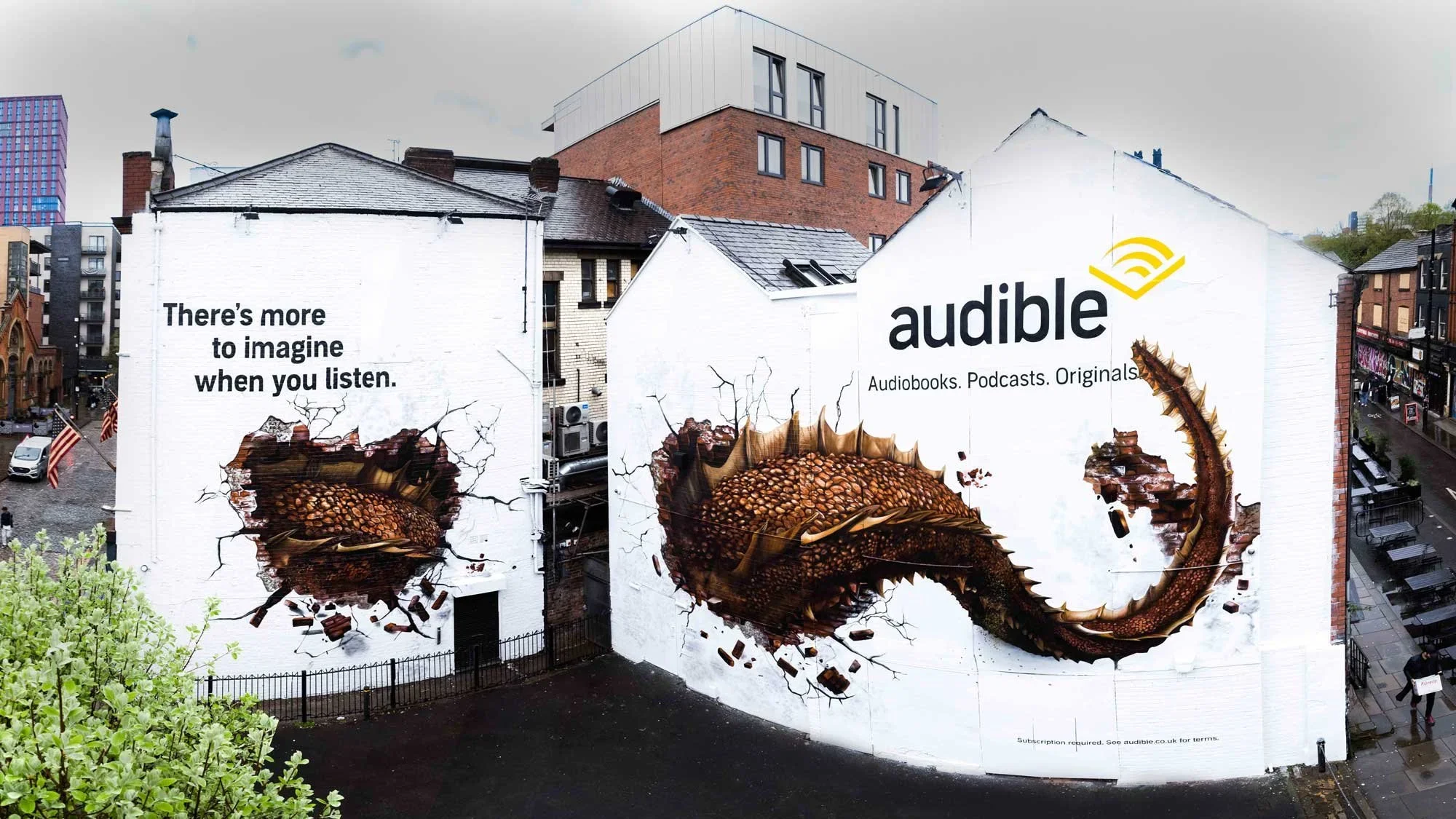 A large outdoor mural advertisement for Audible features a giant animated dragon bursting through the wall, with the dragon's tail and head extending beyond the mural. The mural includes text that reads, 'There's more to imagine when you listen,' and