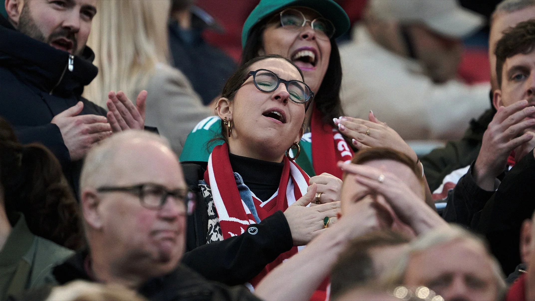 A group of people sitting in stadium seats, cheering and clapping, with some wearing scarves and hats, indicating they are attending a sports event.