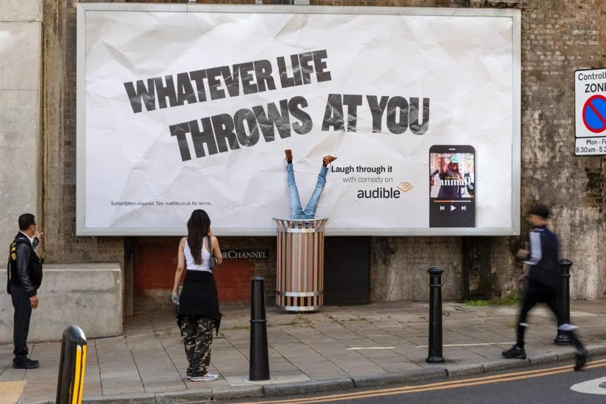 A street scene with a large billboard featuring the slogan 'Whatever life throws at you,' with a person doing a handstand in front of it, fitting their legs into a trash can. Three people are walking by on the sidewalk.