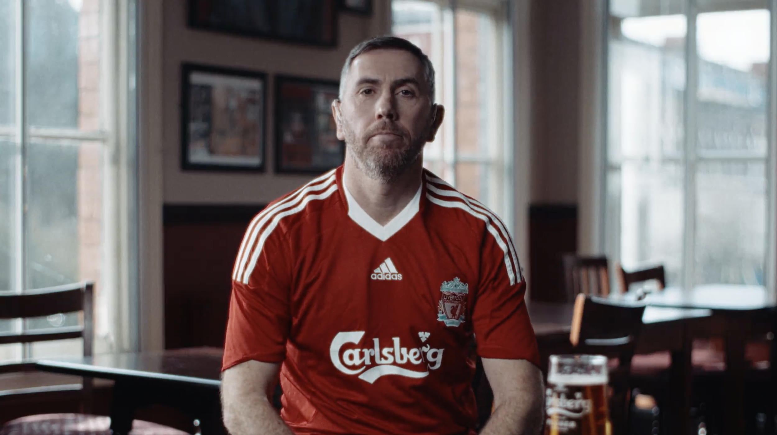 A middle-aged man with a beard and short hair wearing a red Liverpool football jersey sitting indoors at a restaurant or cafe.