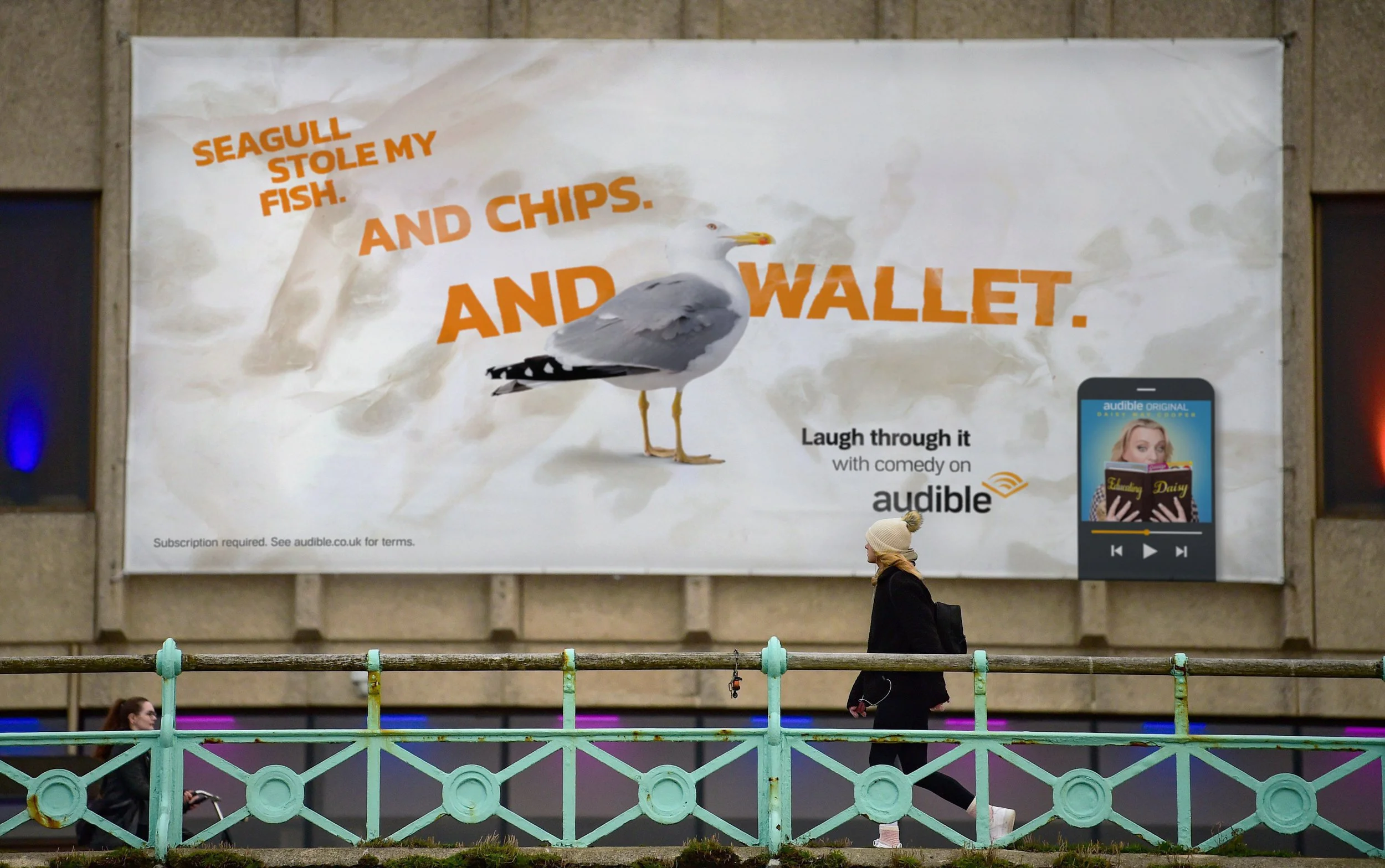 Large billboard advertising Audible featuring a seagull cartoon and text that says 'Seagull stole my fish and chips and wallet.' A person wearing a beige hat and black coat walks past in front of the billboard, with other pedestrians visible below.