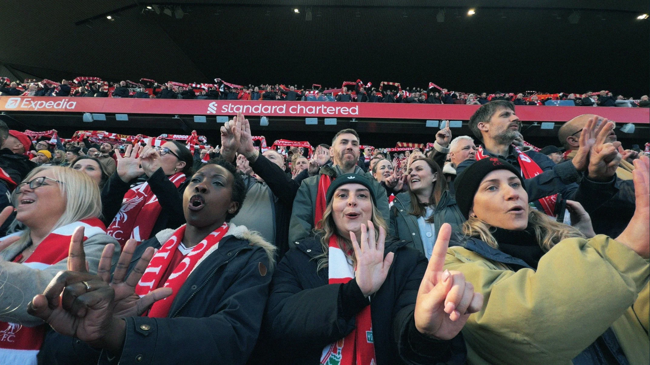 Crowd of enthusiastic soccer fans wearing red scarves and jackets, cheering inside a stadium, with banners displaying advertisements.