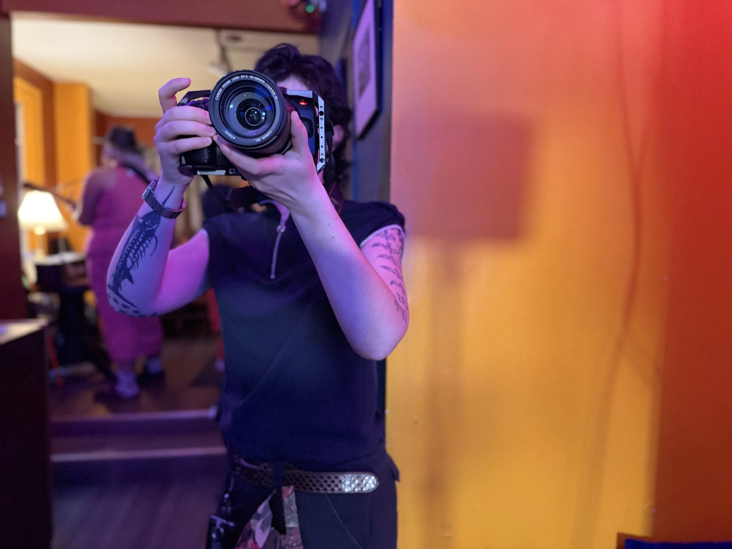 Person taking a photo with a camera reflected in a mirror, tattoos on their arms, wearing black clothing, in a room with vibrant colors.