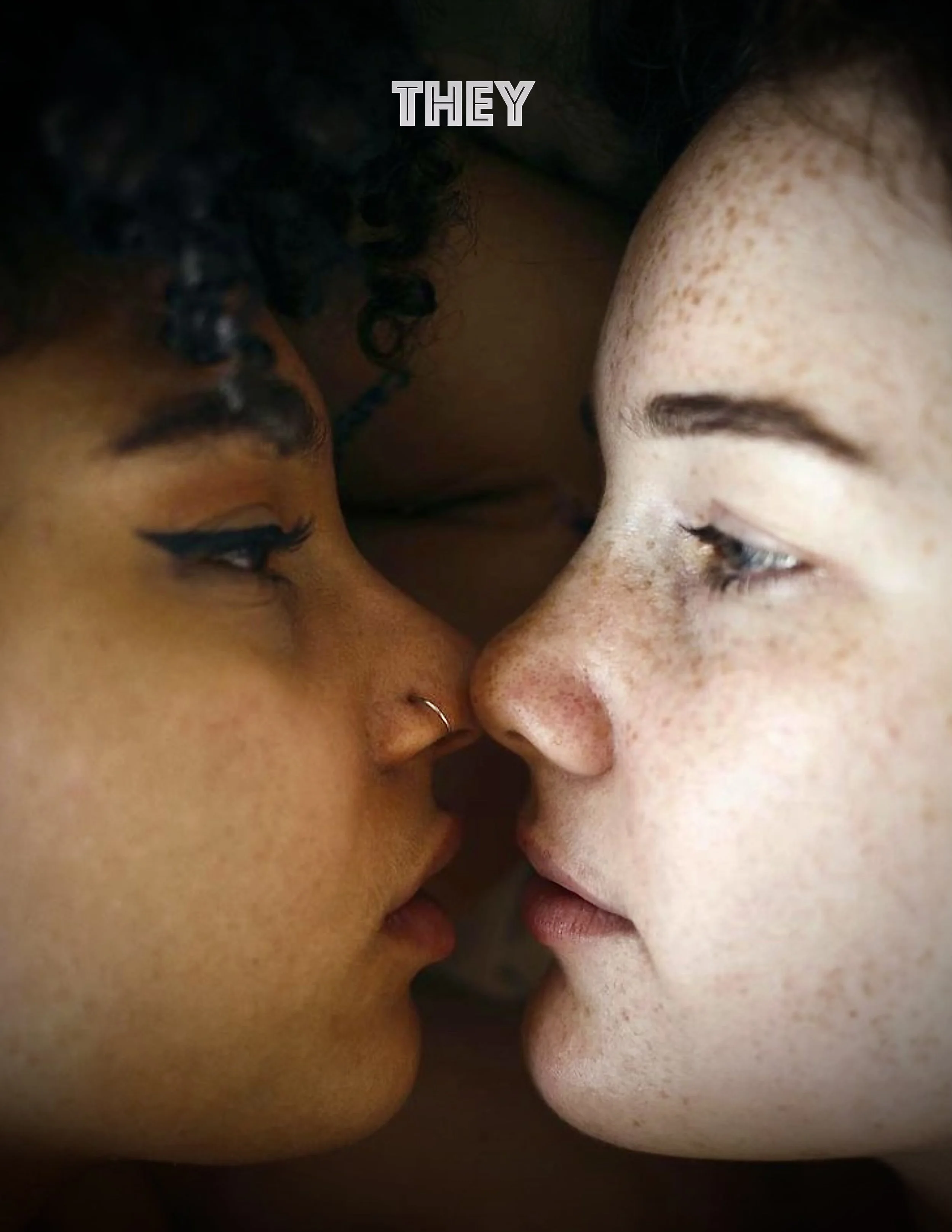 Close-up of two faces touching, one with dark curly hair and freckles, and the other with light skin and freckles, with the word 'THEY' at the top.