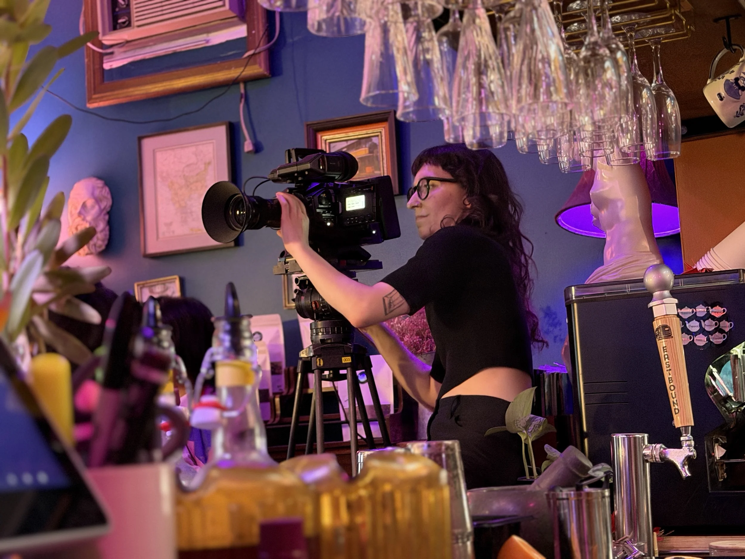 A person with glasses operating a professional video camera in a colorful, decorated room with glasses hanging upside down overhead, framed pictures, and various party supplies.