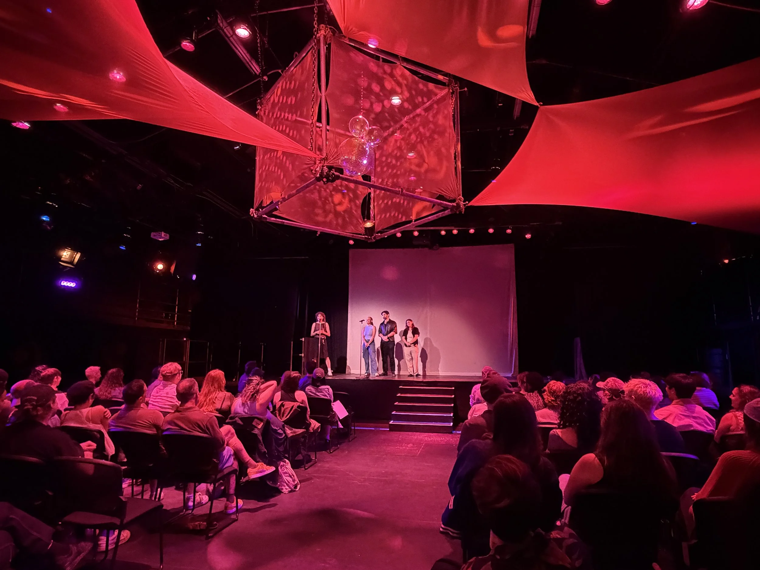 Stage with four speakers talking to audience, large red fabric structures overhead, disco ball, and stage lighting in a dark theater.