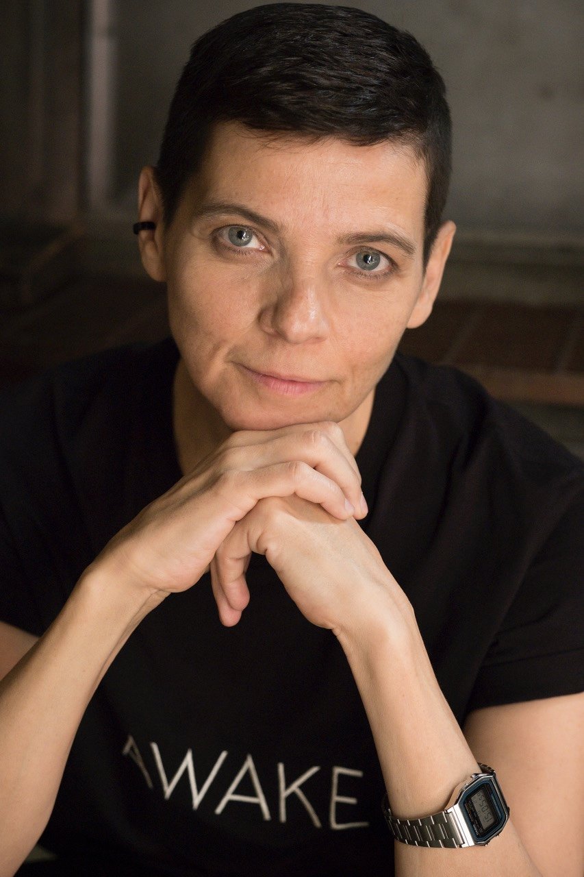 A person with short dark hair, sitting with their chin resting on their clasped hands, wearing a black T-shirt with partially visible white text and a silver wrist watch, looking directly at the camera with a neutral expression.