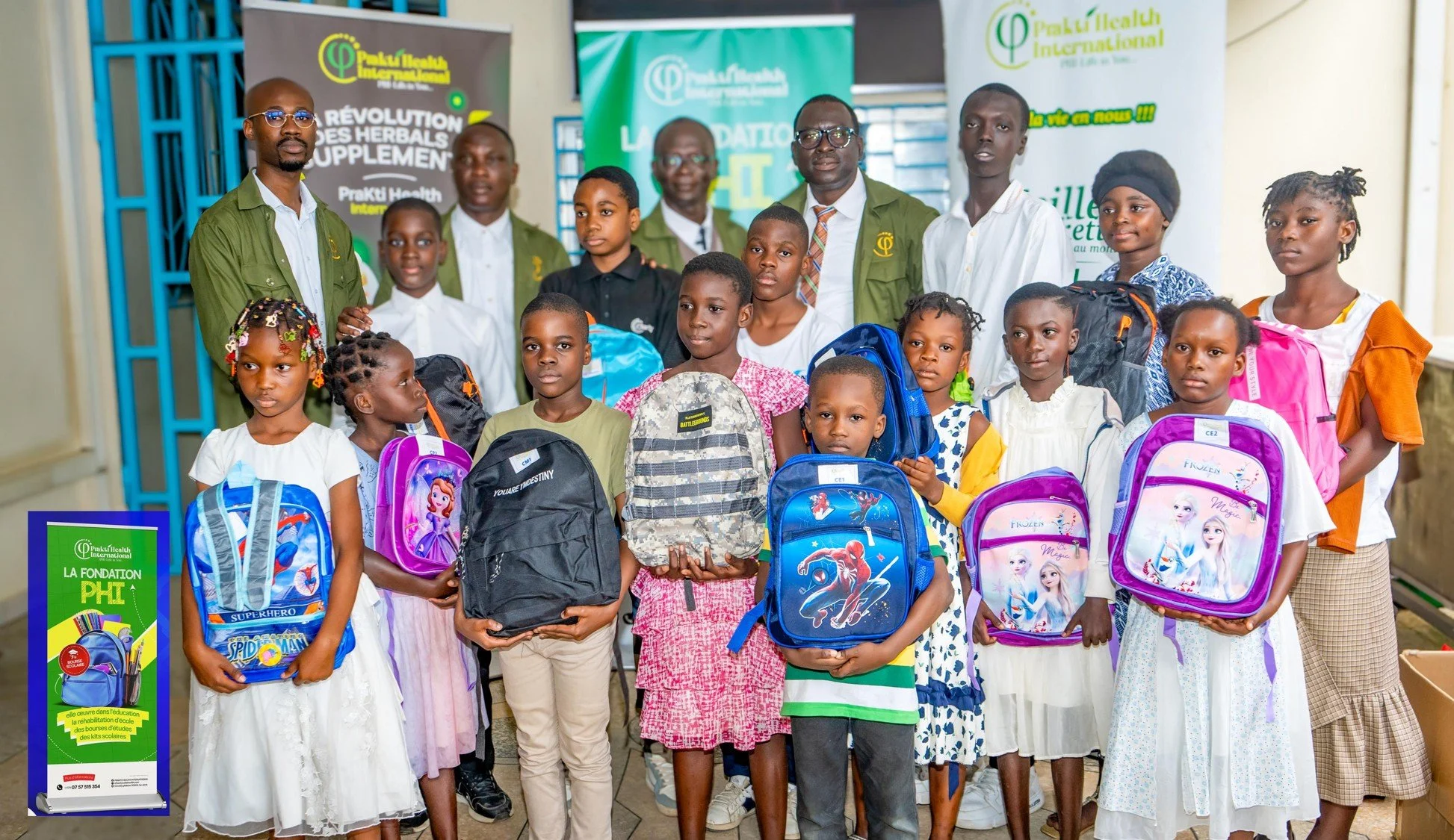 Groupe d'enfants et d'adultes lors d'une cérémonie de distribution de sacs à dos à l'intérieur d'un bâtiment, avec des affiches promotionnelles en arrière-plan. Les enfants tiennent leurs nouveaux sacs à dos aux motifs de personnages de dessins animés.