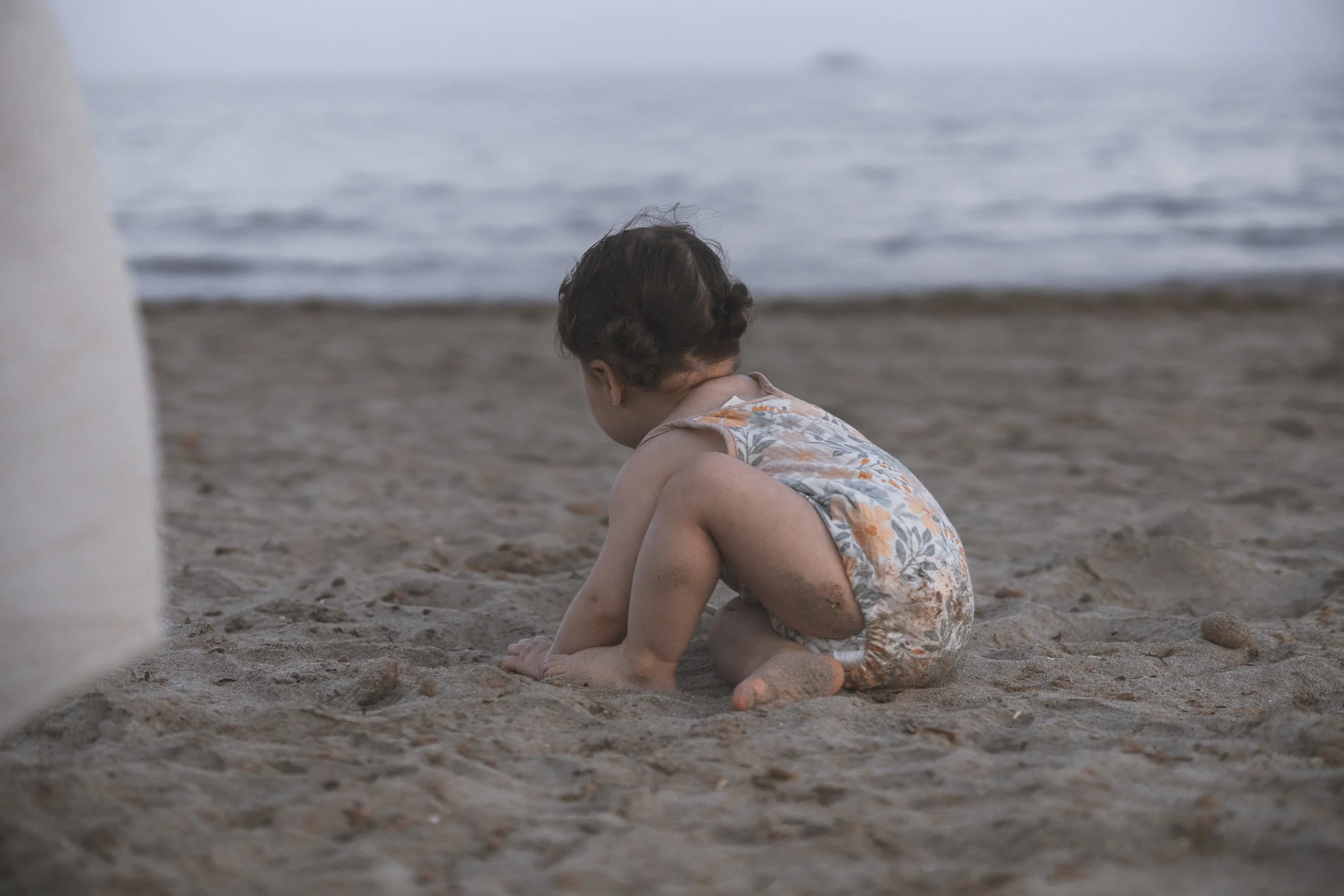 Niña sentada en la playa con cabello rizado, vestida con un vestido de flores, parece estar jugando con la arena frente al mar con olas suaves.