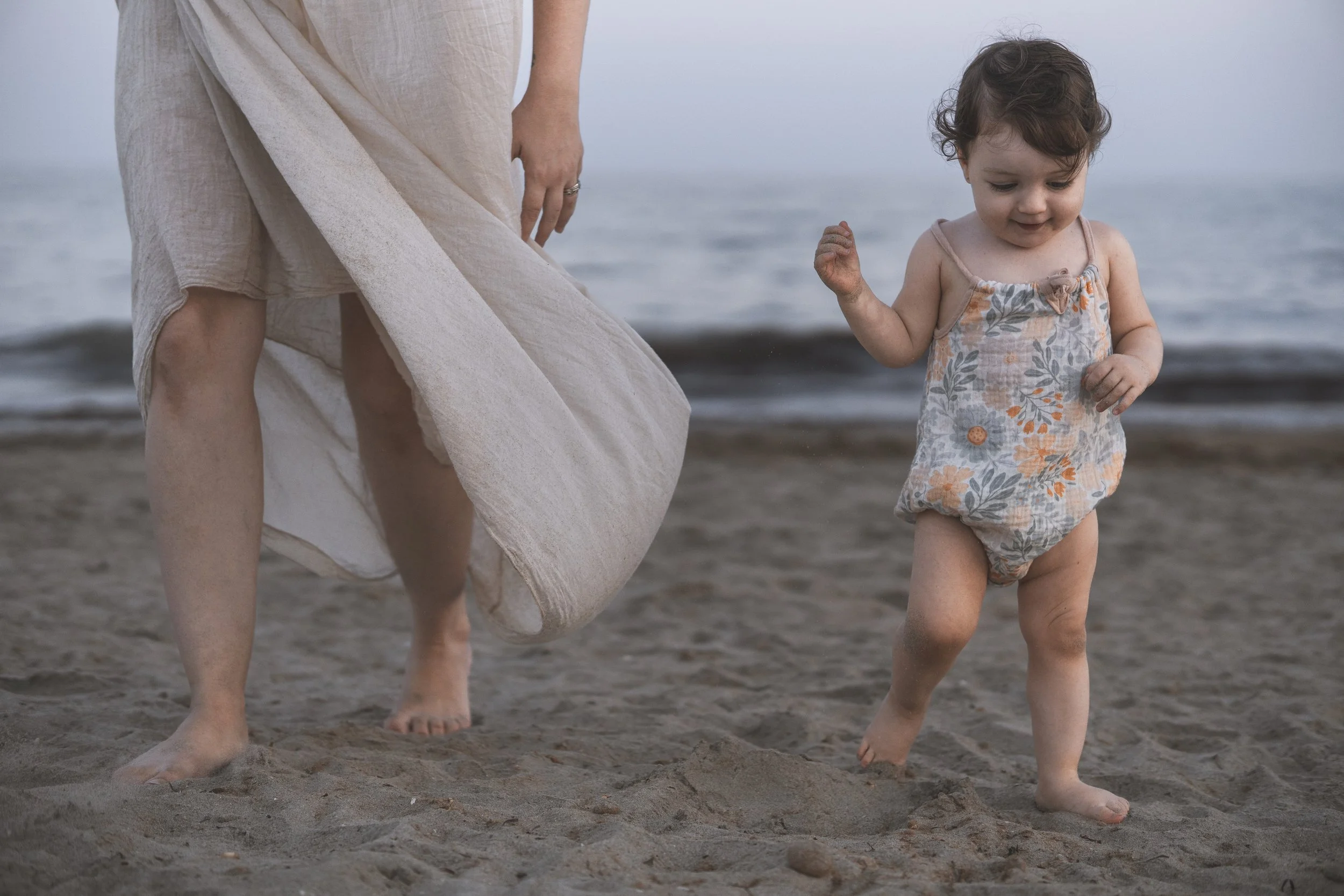 Una niña pequeña con vestido de colores pastel caminando sobre la arena en la playa, acompañada por un adulto que la sostiene de la mano, con el mar de fondo.