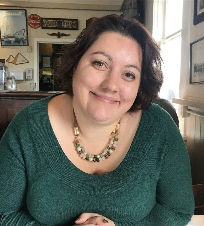 A woman with short dark hair smiling at the camera, wearing a green sweater and a beaded necklace, sitting in a cozy restaurant or cafe with framed pictures and vintage signs on the wall in the background.