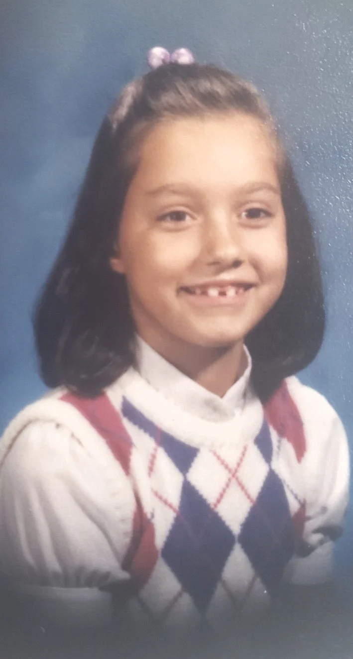 Smiling young girl with dark hair, wearing a white sweater with pink, white, and purple argyle pattern, posed against a blue background.
