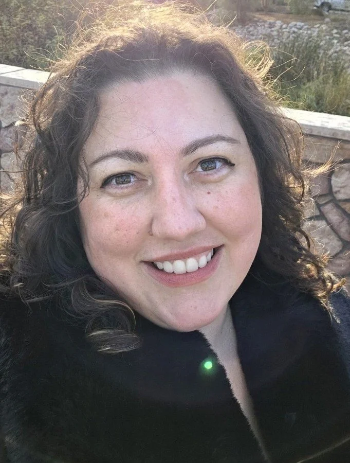 A woman with curly brown hair smiling outdoors, wearing a black coat, with a stone wall in the background.