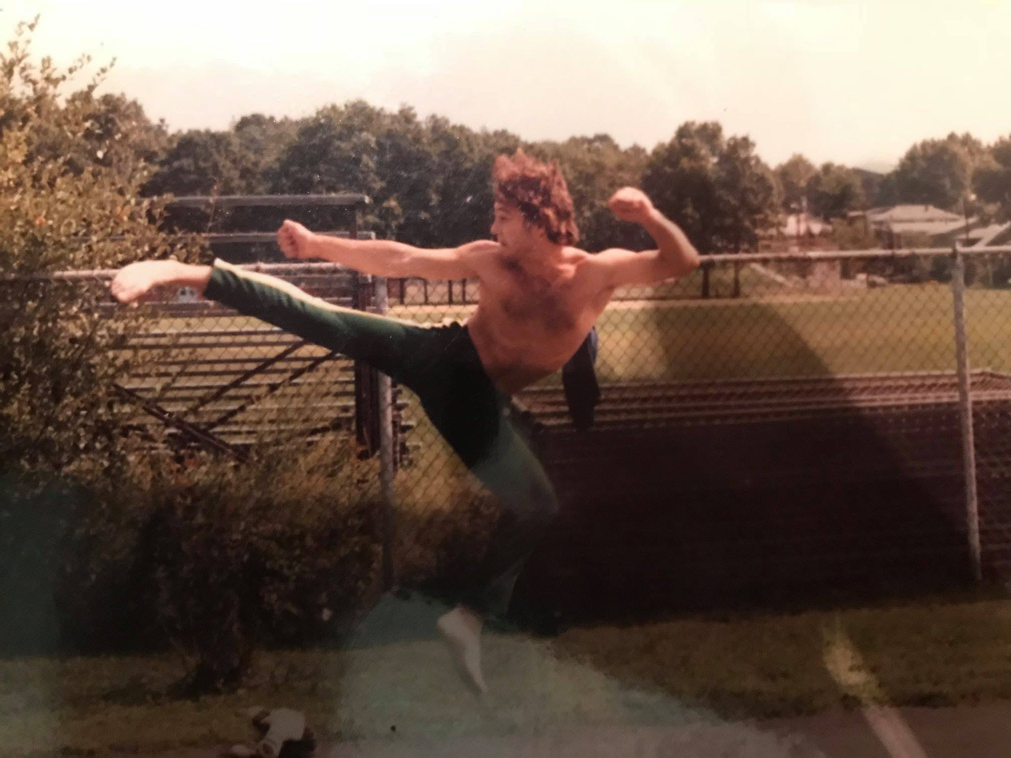 A shirtless person with curly hair performing a high kick in an outdoor fenced area during daytime.