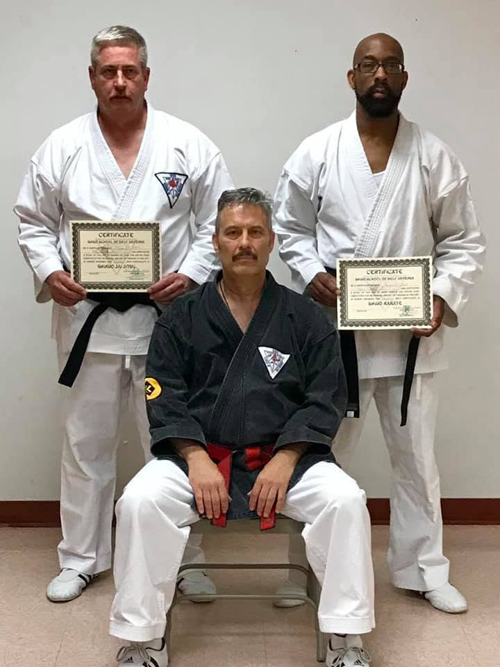 Three men dressed in martial arts uniforms, with two standing and holding certificates, and one sitting in front. The seated man wears a black uniform, while the two standing men wear white uniforms.