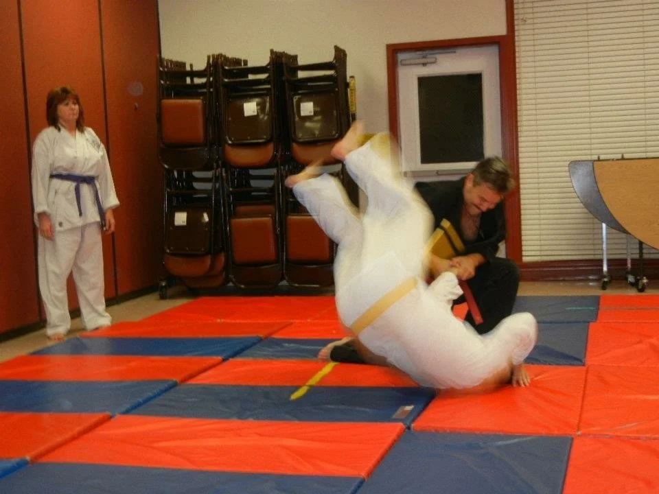 Two people practicing judo or Brazilian jiu-jitsu on mats with a woman in a white gi and a purple belt and a man in a black gi, during a training or tournament session, with others observing in the background.