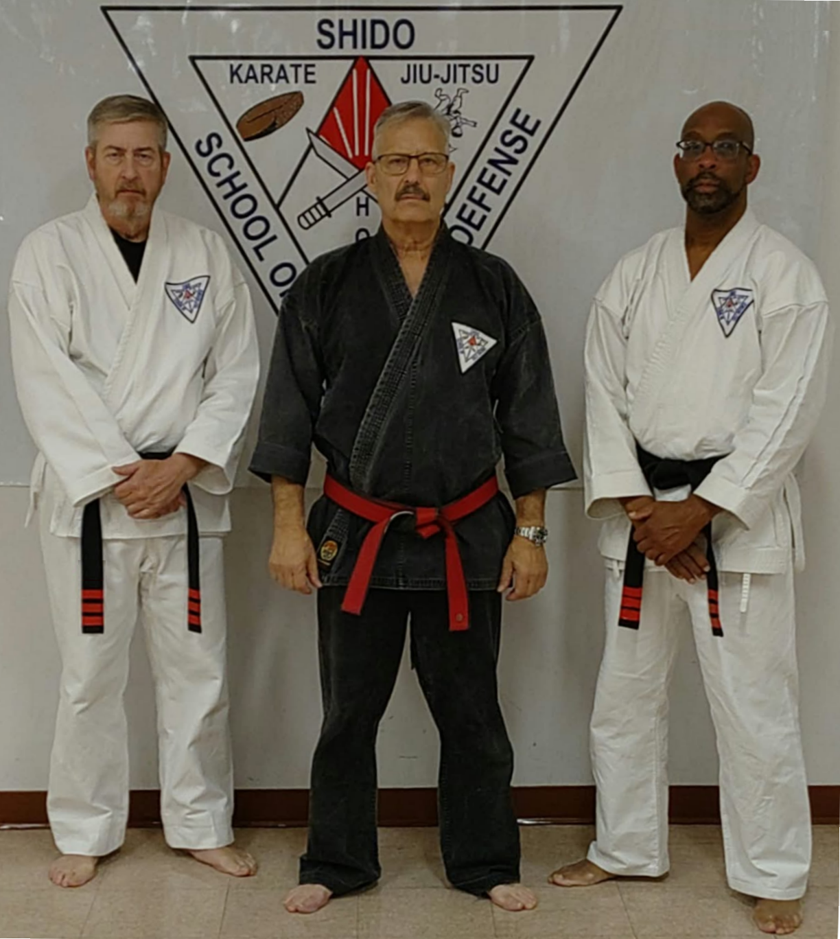 Three martial artists in gi uniforms standing in front of a martial arts banner with logos and text, two wearing white gis and one in a black gi with a red belt.