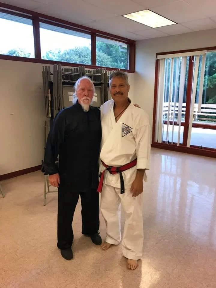 Two men standing together, one in a black outfit and the other in a white martial arts gi with a red and black belt, smiling inside a room with large windows.