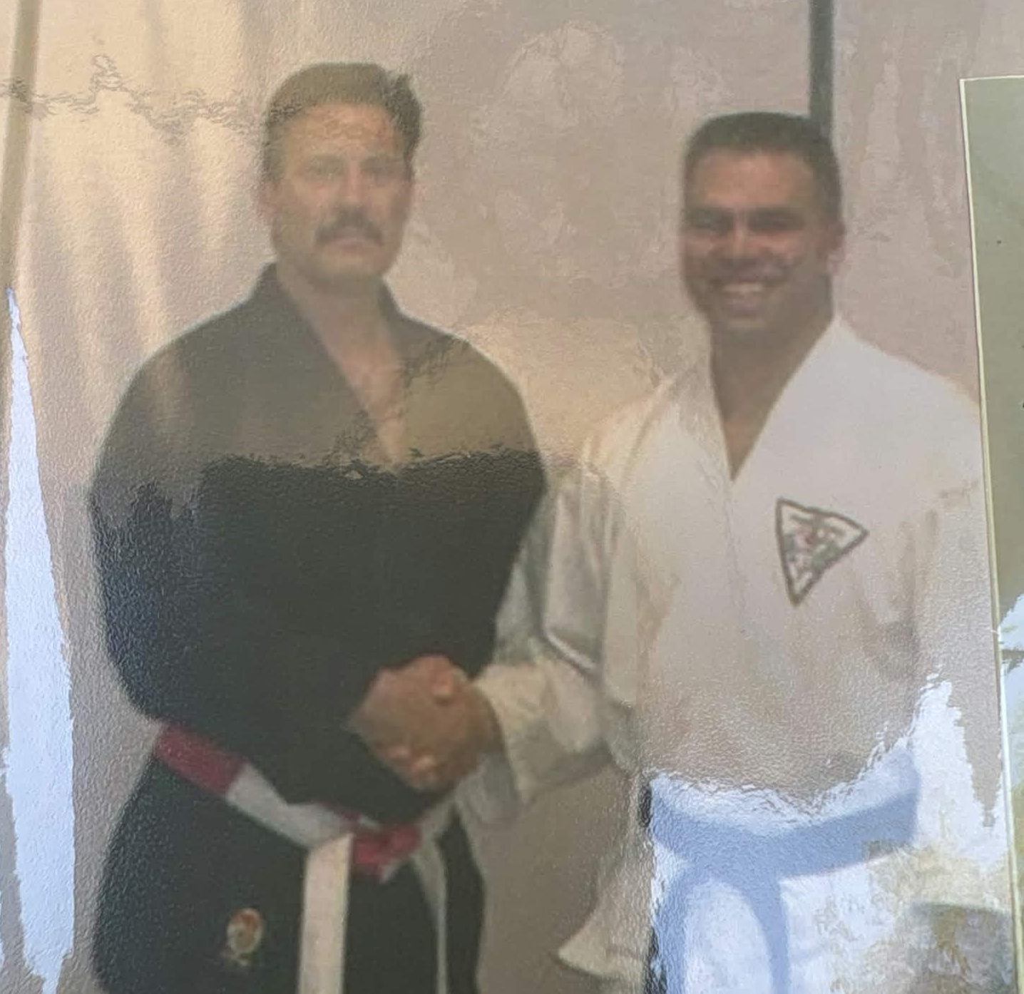 Two men shaking hands, one in a black traditional outfit and the other in a white martial arts gi with a badge, in an indoor setting.