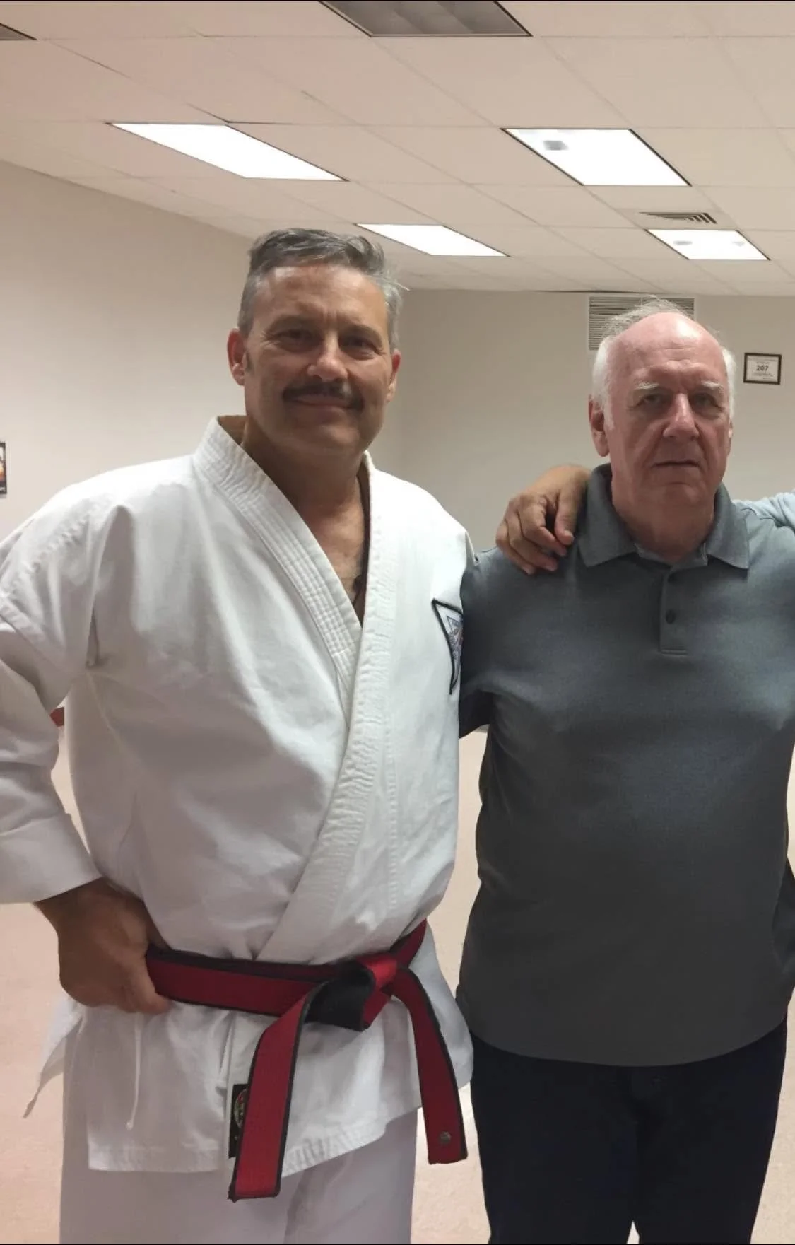 Two men standing side by side in a room; one is wearing a karate uniform with a red belt, and the other is in a gray polo shirt. The man in karate has his arm around the other man's shoulder, and they are both smiling.