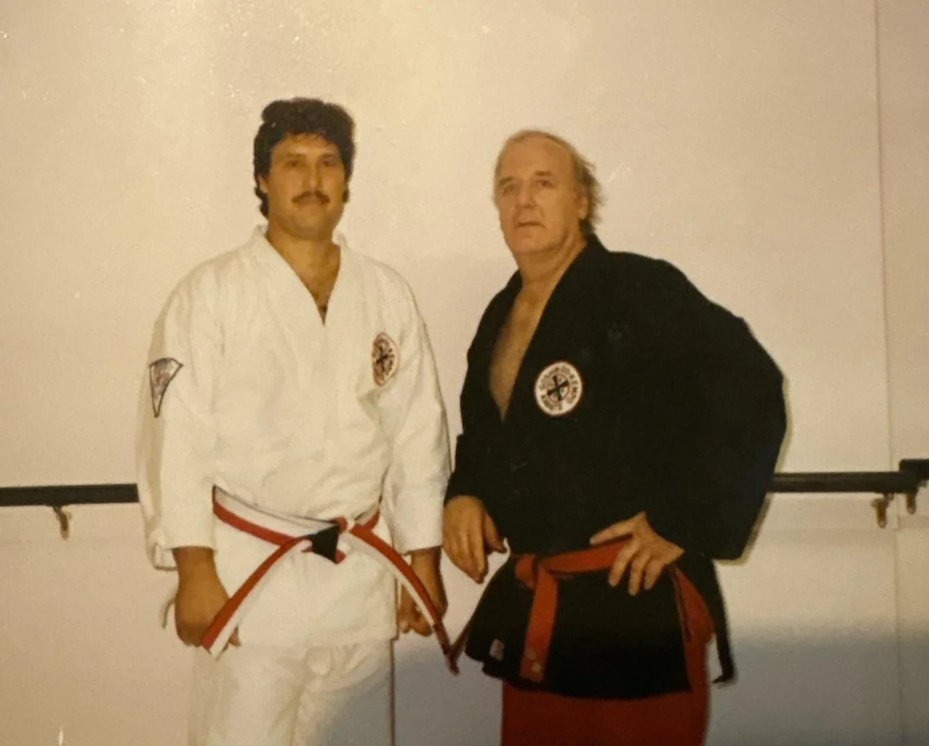 Two men in martial arts uniforms posing indoors, with a white wall and a black horizontal bar behind them.
