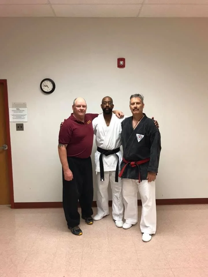Three men standing together in a room, with two dressed in martial arts uniforms and one in a casual polo shirt. The room has beige walls, a clock, and a fire alarm visible.