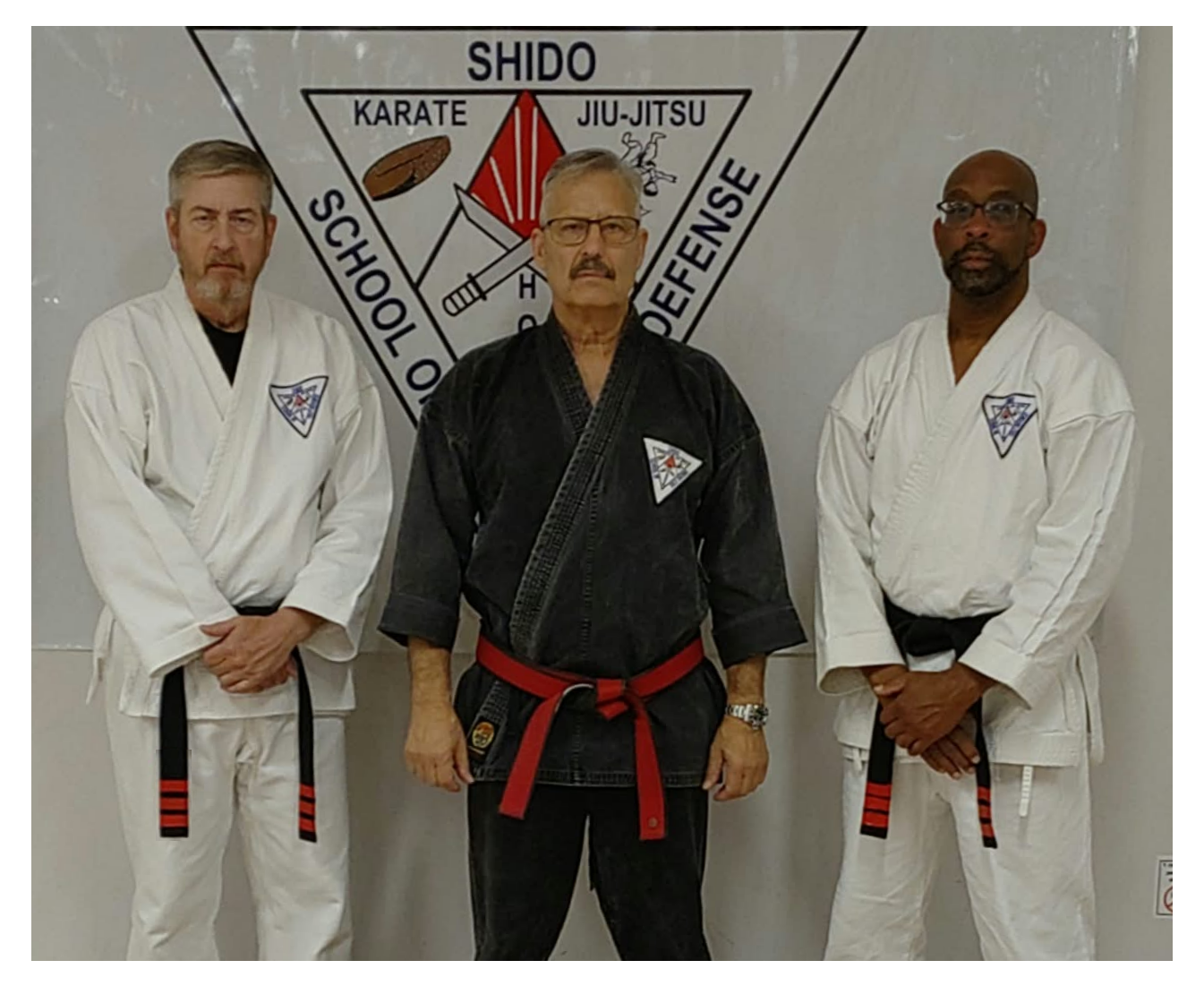 Three men in martial arts uniforms standing in front of a martial arts academy sign. The man in the center wears a black gi with a red belt, and the two on either side are in white gis with black belts. The background features a logo with the words 'School of Protection,' 'Karate,' 'Jiu-Jitsu,' and 'Shido.'