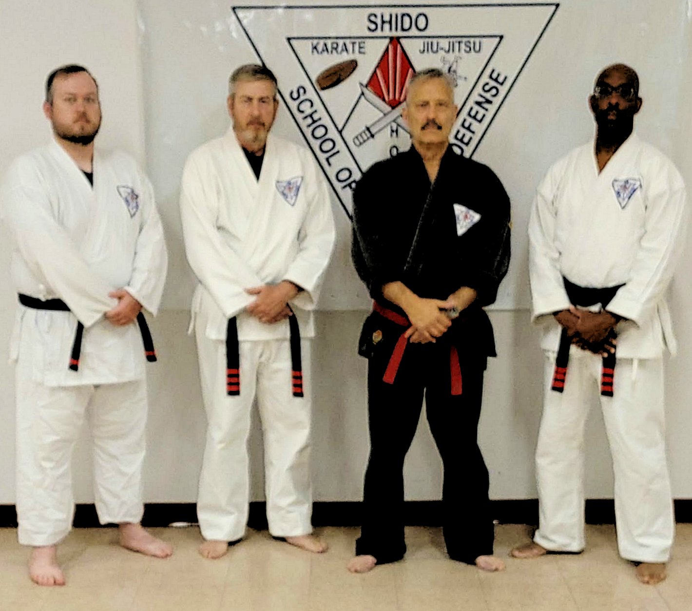 Four men in martial arts uniforms standing in front of a wall with a martial arts emblem. Three are in white gi and one in black gi, all with black belts. The emblem features a triangle with martial arts disciplines and the words "School of" and "Defense."