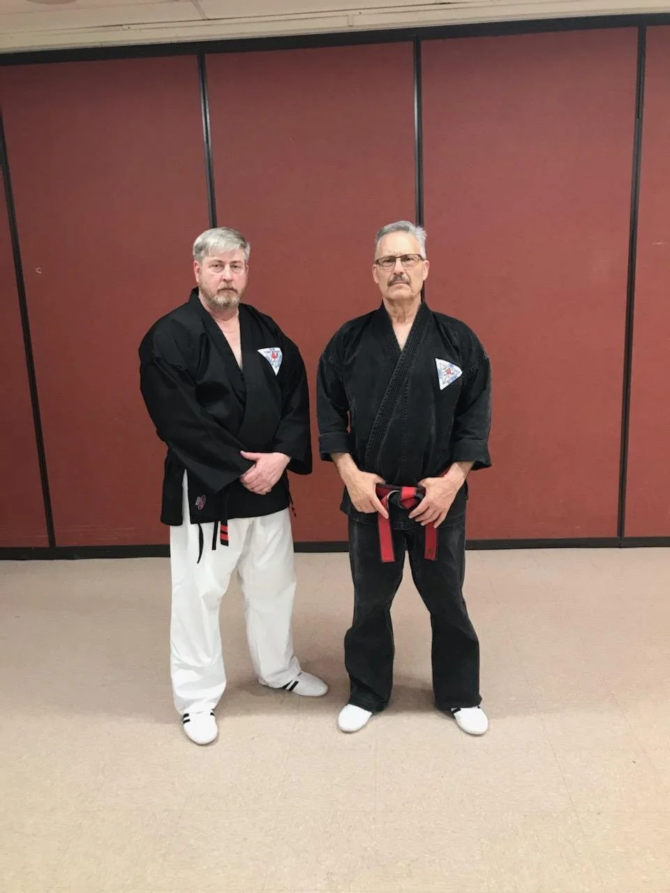 Two men in martial arts uniforms standing in front of a red folding partition. The man on the left has gray hair and a beard, wearing a black top and white pants. The man on the right has gray hair and glasses, wearing a black gi with a black belt.