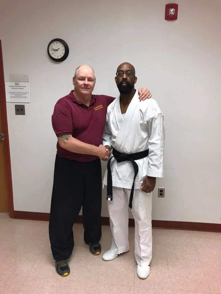 Two men shaking hands, one in a maroon polo shirt and black pants, the other in a white martial arts gi with a black belt, standing in a room with a clock on the wall.