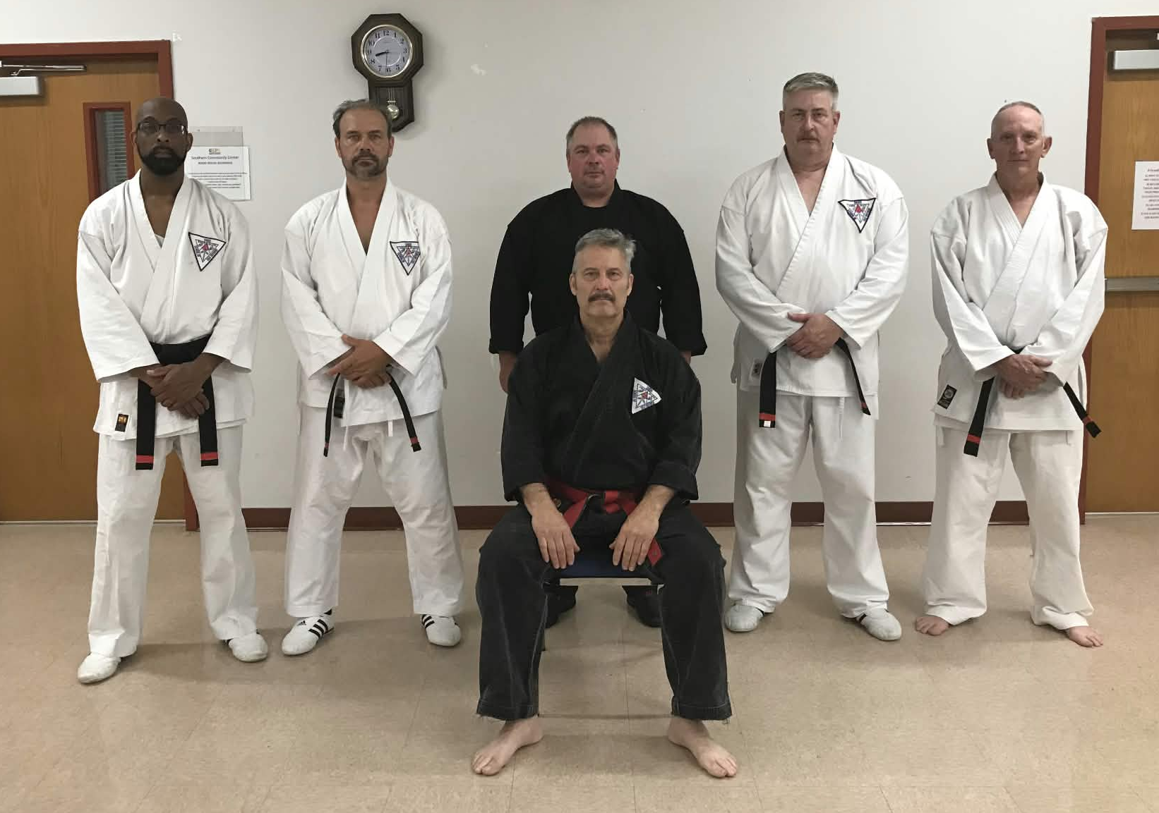 Group of six men in martial arts uniforms in a dojo, with two sitting in front and four standing behind, with a clock on the wall.